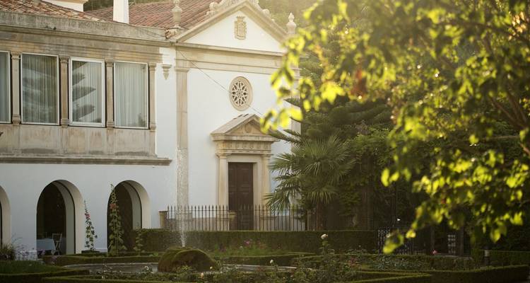 Jardín salpicado de sol y fachada de villa histórica con ventanas arqueadas y detalles ornamentales.