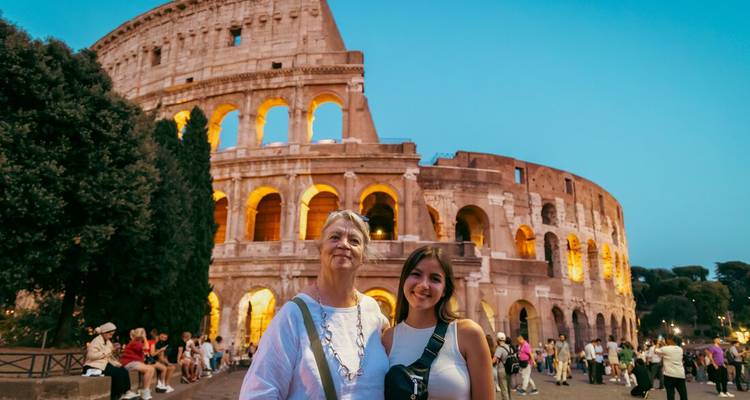 Mujer mayor y menor sonriendo frente al Coliseo iluminado de Roma al anochecer.