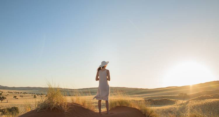 Woman in dress and sunhat gazes over sunlit savanna from the crest of a reddish dune at sunrise