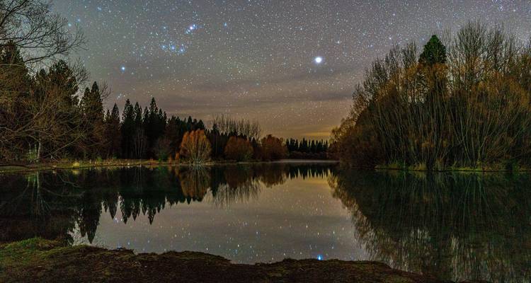 Cielo nocturno lleno de estrellas que se refleja en un lago tranquilo del bosque rodeado de árboles otoñales.
