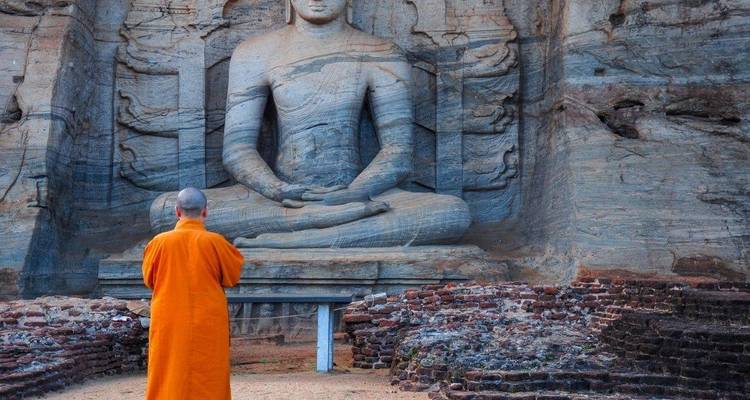 Monje con túnica naranja frente a una gran talla de Buda en roca.