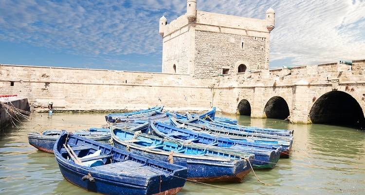 Blick auf blaue Fischerboote in einem Hafen mit einer Steinfestung.