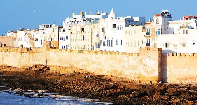 Küstenblick auf weiße Gebäude vor einem blauen Himmel mit einer alten Mauer und felsiger Küstenlinie.
