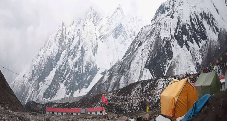 Colorful tent camp and prayer flags sit below steep snow-covered Himalayan peaks beneath cloudy skies.