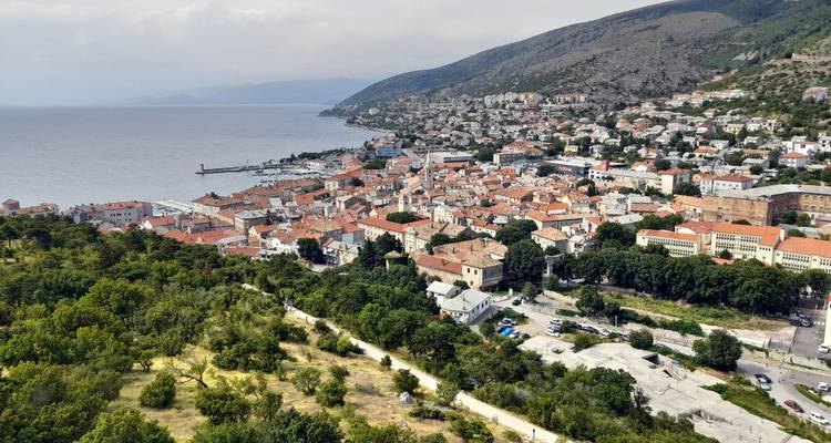 Vue aérienne d'une ville côtière aux toits rouges s'étendant le long d'une péninsule avec des collines environnantes et la mer.