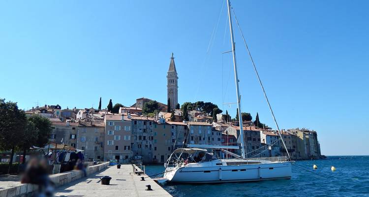 Segelboot, das vor der pastellfarbenen Hafenfront und dem hohen Glockenturm der Altstadt von Rovinj unter klarem Himmel angelegt hat.