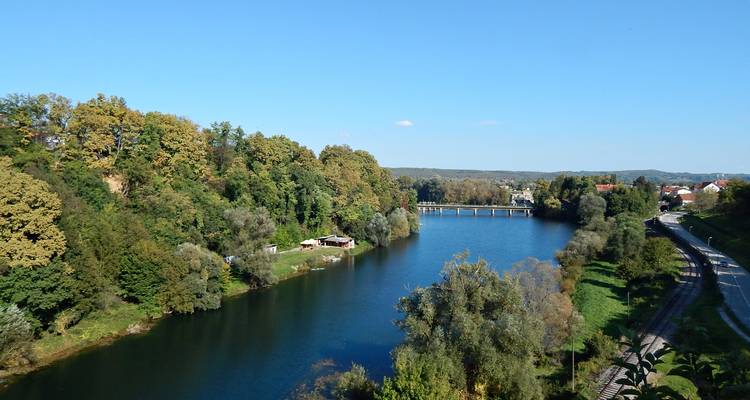 Serene rivier omgeven door dicht groen bos onder een helderblauwe lucht met een klein bruggetje in de verte.