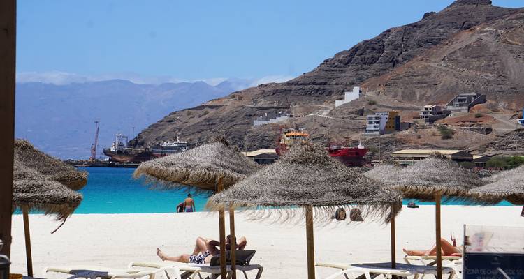 Strandszene mit Menschen unter Stromsonnenschirmen und entfernten Schiffen.