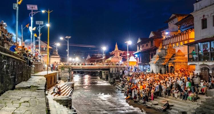 Crowded riverside scene in the evening with illuminated structures and a bridge.