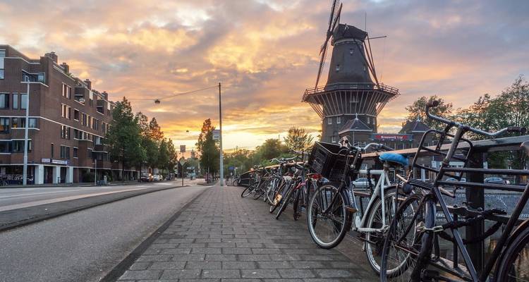 Row of bicycles parked beside a canal with a traditional Dutch windmill at sunset in Amsterdam