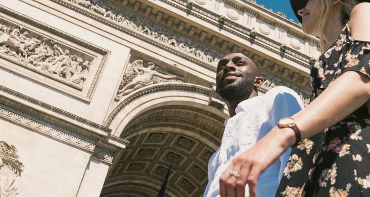 Upward angle of couple walking beneath the Arc de Triomphe’s ornate stonework on a bright day.