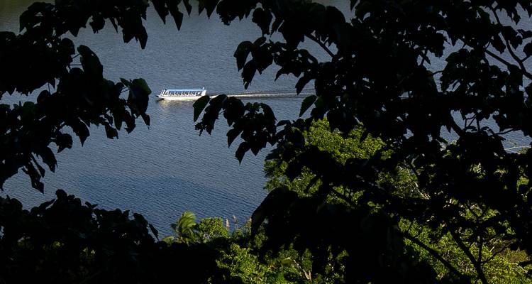 Croisière en bateau long sur un lac bleu encadré par des feuillages sombres au premier plan créant une vignetted naturelle.