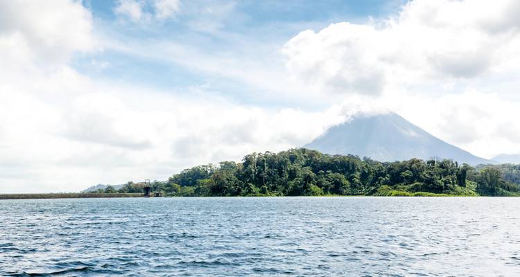 Lac calme avec une île forestière et le cône du volcan Arenal partiellement voilé par les nuages.