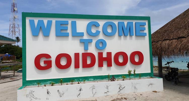 Letrero en negrita al costado del camino que dice Bienvenido a Goidhoo en un día soleado en la isla