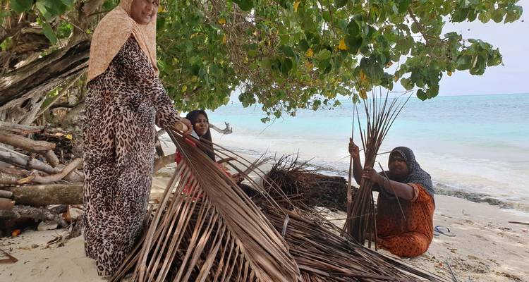 Mujeres locales en una playa arenosa tejiendo frondas de palma bajo la sombra de árboles frondosos y el mar turquesa