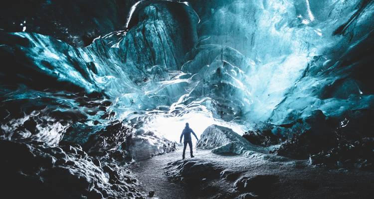 Une silhouette se dresse à l'entrée brillante d'une immense grotte de glace turquoise.