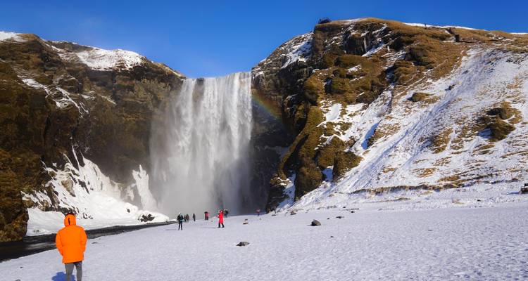 Rainbow arcs across Skogafoss waterfall surrounded by snowy landscape and visitors in winter gear.