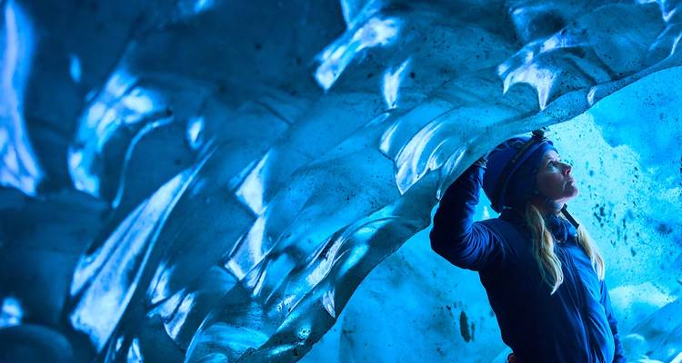 Woman gazes up at shimmering blue ice formations inside a glacier cave.