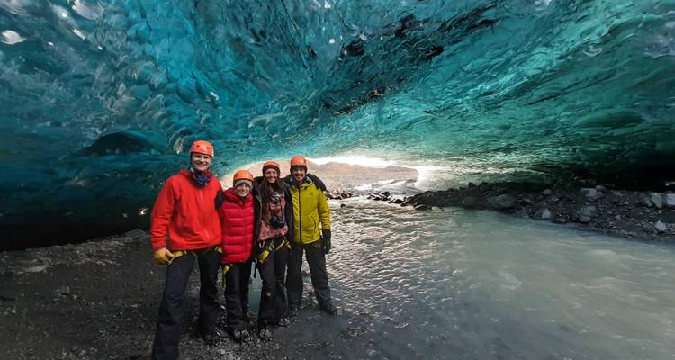 Smiling group poses inside a brilliant blue ice cave beside a glacial river.