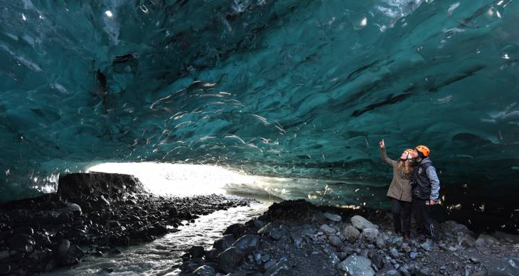 Couple explores a turquoise ice cave illuminated by natural light at its opening.