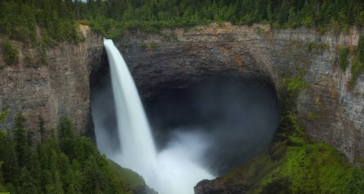 Powerful waterfall plunges into a mist-filled amphitheatre of dark basalt cliffs surrounded by dense conifer forest.