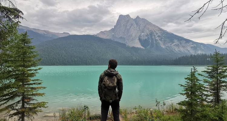 Ein einzelner Wanderer mit einem Rucksack blickt über einen türkisfarbenen Alpensee auf bewaldete Berge und einen gezackten Gipfel unter einem bewölkten Himmel.