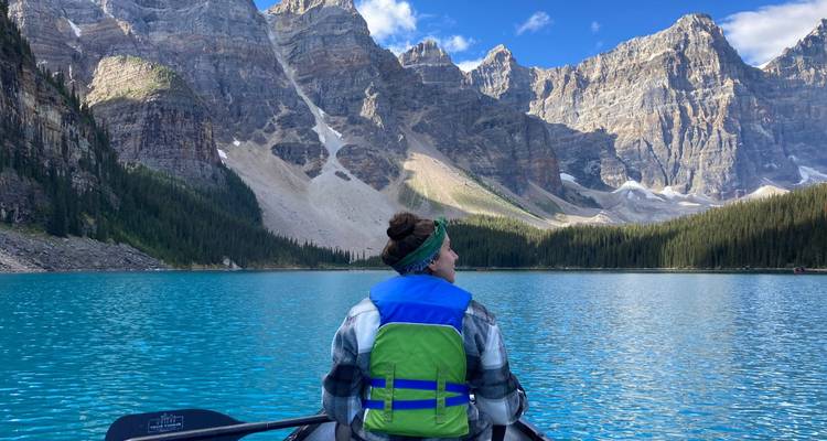 Eine Frau in einer Schwimmweste sitzt in einem Kanu auf einem strahlend blauen Bergsee und blickt auf hohe felsige Gipfel.