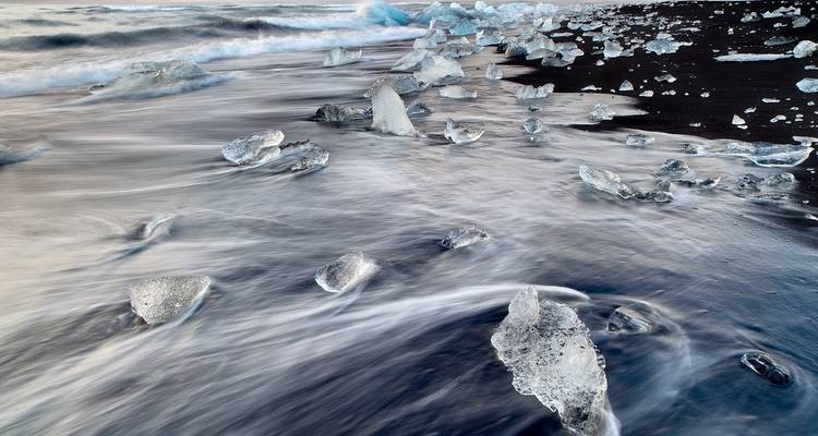 Des blocs de glace transparente sont dispersés sur un rivage de sable noir tandis que des vagues soyeuses se déploient sur la plage.