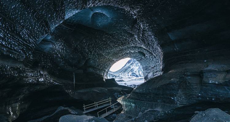 À l'intérieur d'une grotte de glace bleue scintillante, une ouverture circulaire révèle des pics enneigés et une passerelle artificielle en dessous.