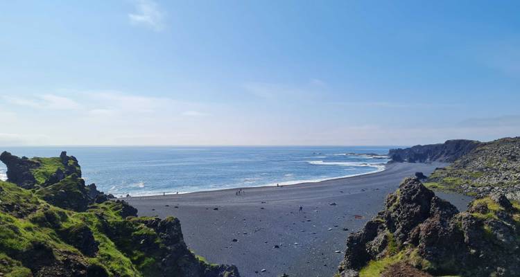 Vaste plage de galets noirs bordée par des falaises volcaniques dentelées et l'océan bleu éclatant.