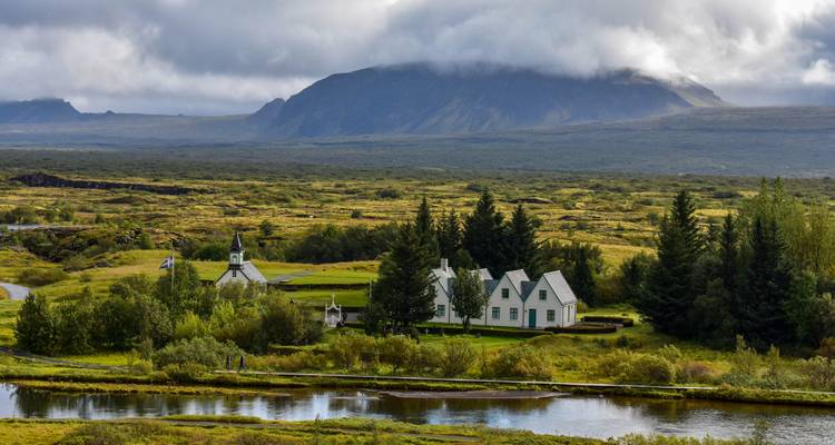 L'église blanche historique et les maisons de Thingvellir sont situées à côté d'une rivière avec des montagnes brumeuses au-delà.