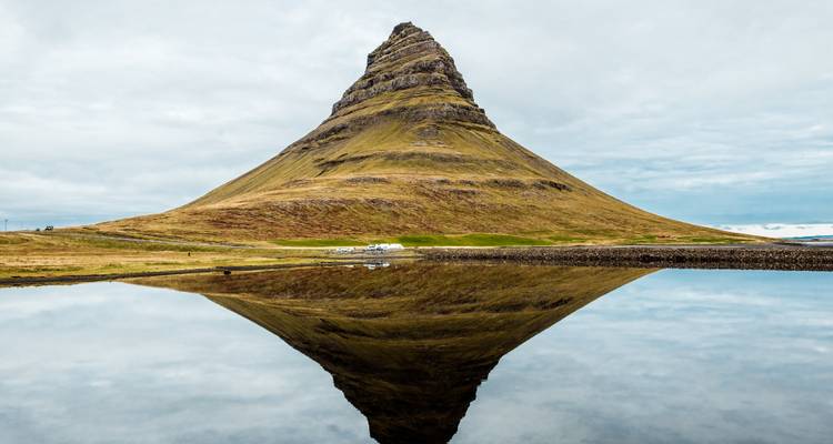 Le cône distinct de la montagne Kirkjufell s'élève au-dessus d'une eau calme, parfaitement reflété dans son reflet par un jour nuageux.