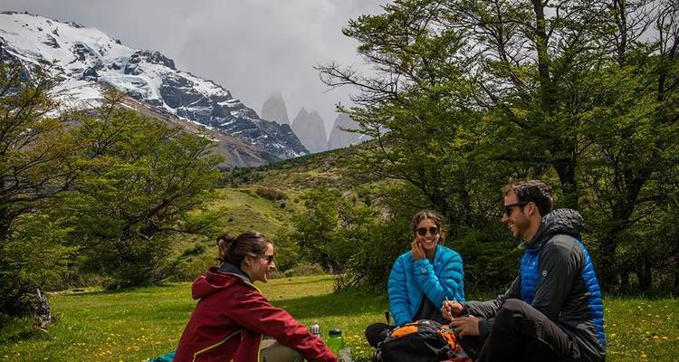 Drie trekkingstoeristen zitten op een grazig weiland te genieten van een picknick met met sneeuw bedekte graniettorens die boven groene heuvels uitrijzen.