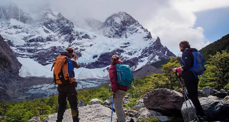 Wandelaars met rugzakken kijken omhoog naar een imposante gletsjer en met sneeuw bedekte bergtoppen onder dramatische wolken.