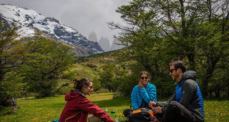 Drie trekkers ontspannen op het gras en genieten van hun lunch terwijl de iconische Torres del Paine pieken imposant in de mist achter hen uitsteken.