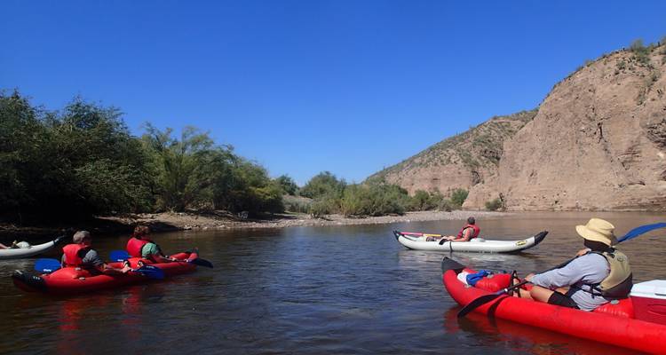People kayaking on a calm river with rocky banks.