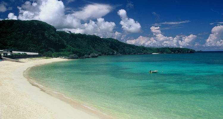 Sandy beach with clear azure waters, and green-covered hills under a bright sky.