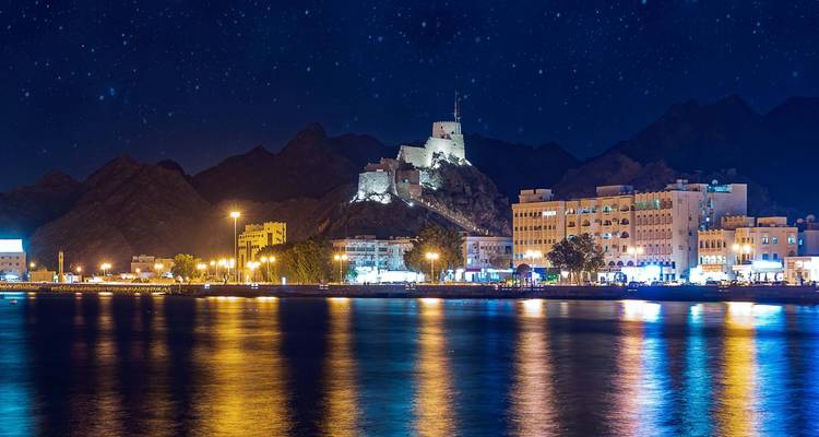 Paisaje urbano nocturno reflejándose hermosamente en un cuerpo de agua con montañas al fondo.
