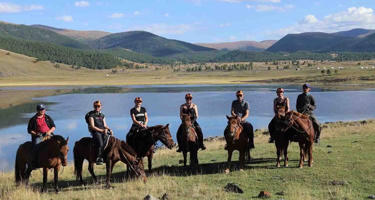 Grupo de personas a caballo cerca de un lago con montañas al fondo.