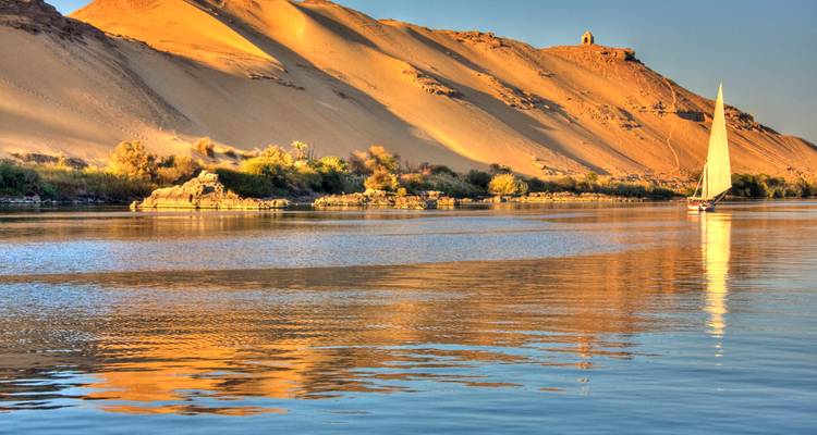 Un velero solitario se desliza en el reflectivo río Nilo junto a las doradas dunas del Sahara al atardecer.
