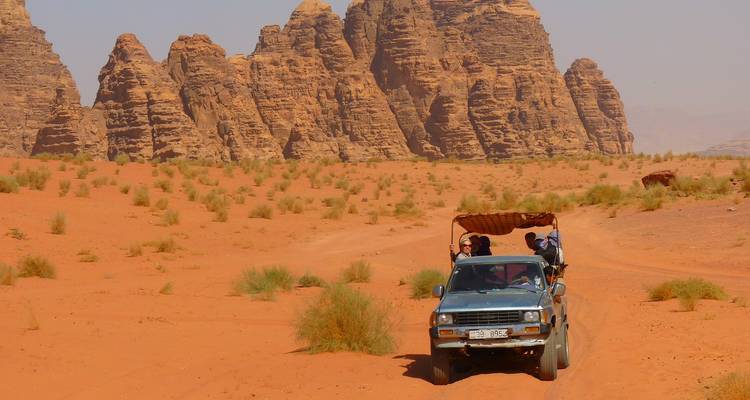 Ein offenes Jeep-Fahrzeug mit Touristen fährt über die rötliche Wüste mit aufragenden Felsformationen des Wadi Rum.