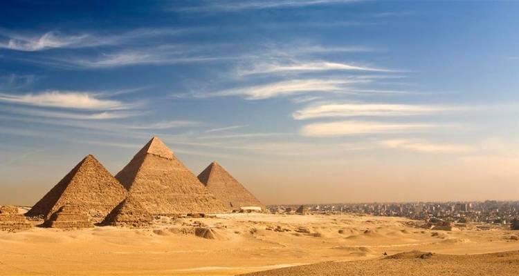 Three pyramids rise from expansive golden desert sands beneath wispy clouds on a clear day.