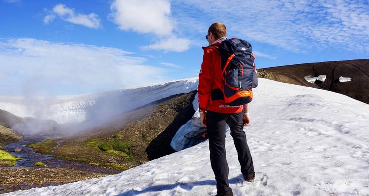 Un excursionista con chaqueta roja camina por una meseta volcánica nevada junto a un borde de hielo humeante bajo un cielo azul.
