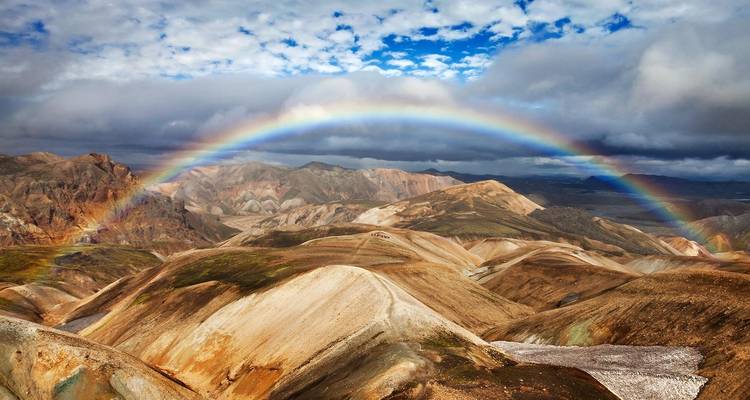 Un arco iris brillante arqueándose sobre colinas de riolita multicolor y accidentadas bajo un cielo parcialmente nublado.