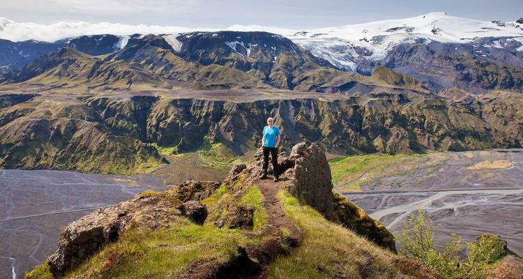 Vista trasera de un trekker de pie en una cresta cubierta de pasto con un cañón dramático y montañas coronadas de glaciares más allá.