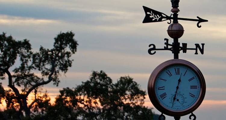 Veleta brújula decorativa con reloj de números romanos contra un cielo al atardecer lleno de colores