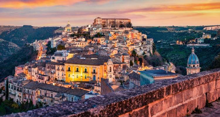 Farbenfrohe Ansicht einer Hügelstadt bei Sonnenuntergang, mit historischen Gebäuden und einer malerischen Landschaft.