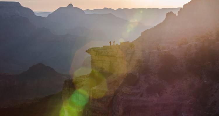 Deux petites silhouettes se tiennent debout sur un affleurement rocheux d'un canyon au lever du soleil avec des reflets de lumière.