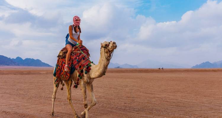 Eine Person, die in der Wüste auf einem Kamel reitet, mit Bergen im Hintergrund.