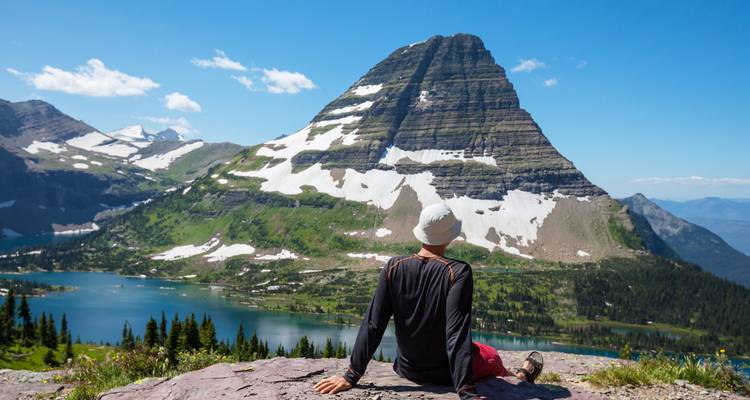 Traveler in a white sunhat sits on a rock overlooking Hidden Lake and Bearhat Mountain.